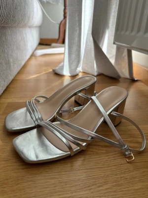 Silvriga klackar med öppen tå - Använda en gång!🤍 Säljer ett par stilrena sandaletter i metallic silver med smala remmar och fyrkantig tå. Skorna har en bekväm blockklack och justerbart spänne vid ankeln. Materialet är glansigt och ger en trendig look, perfekt för dig som gillar att sticka ut.