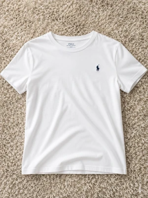 Vit t-shirt från Polo Ralph Lauren - Klassisk vit t-shirt från Polo Ralph Lauren med rund halsringning och korta ärmar. Diskret mörkblå logga broderad på bröstet. Perfekt basplagg i mjuk bomull som passar till allt.