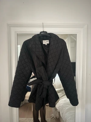 Svart quiltad trenchcoat från NA-KD - Stilren svart quiltad trenchcoat från NA-KD med bälte i midjan och bred krage. Jackan har ett klassiskt rutmönster och är tillverkad i ett mjukt, vadderat material som ger en trendig look. Perfekt för lager-på-lager och höstväder. Mycket fint stick och använd fåtal gånger 💋