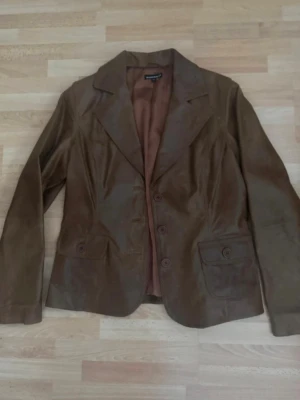 Brun skinnjacka med knappar - Snygg brun skinnjacka med klassisk krage och tre knappar framtill. Jackan har två fickor med lock och knapp, samt en figurnära passform. Perfekt för dig som vill ha en stilren och tidlös look. Storlek Xl, men förminskad som M, bröst 100cm.