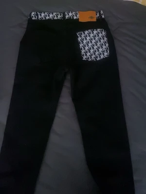 Svarta jeans med monogramdetaljer - Säljer ett par svarta jeans med vita monogramdetaljer i midjan och på bakfickan. Jeansen har en slim fit-modell och en snygg kontrast mellan det svarta tyget och de mönstrade partierna. Perfekta för dig som gillar streetwear och unika detaljer.