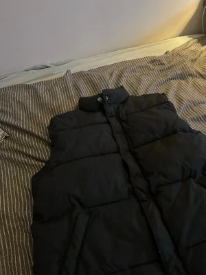 Svart dunväst från Clique - Snygg svart dunväst från Clique med hög krage och quiltad design. Västen har dragkedja och tryckknappar framtill, perfekt för lager på lager under kyliga dagar. Enkel och stilren look som passar till det mesta. Röd logga vid nacken