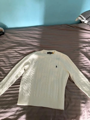 Vit kabelstickad tröja Polo Ralph Lauren - Kabelstickad vit tröja från Polo Ralph Lauren med rund halsringning och broderad logga på bröstet. Tröjan har ribbade muddar vid ärmslut och nederkant, och är tillverkad i mjukt material som ger en klassisk och stilren look.