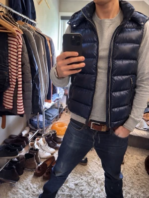 Mörkblå dunväst med hög krage - Snygg mörkblå dunväst med hög krage och dragkedja framtill. Västen har två sidofickor med dragkedja och quiltad design som ger en puffig look. Perfekt att slänga över en hoodie eller tröja för extra värme.