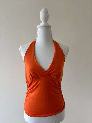 Orange halterneck linnetopp - Snygg orange linnetopp med halterneck och djup v-ringning. Toppen är ärmlös och har en figurnära passform som framhäver siluetten. Perfekt för varma dagar eller som ett statement-plagg i din garderob.