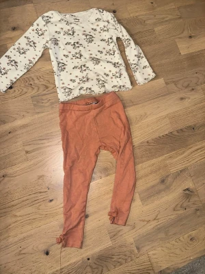 Disney set - Set med en långärmad tröja och ett par byxor. Tröjan är vit med ett mönster av Disney och byxorna är enfärgade i en terrakottafärg med rosetter vid bensluten.