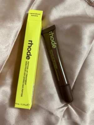 Rhode Peptide Lip Treatment Banana - Rhode Peptide Lip Treatment i smaken Caramelized Banana. Läppbalsamet kommer i en brun tub med gul text och har en smidig applikator. Förpackningen är gul och tydligt märkt med produktnamnet. Innehåller 10 ml.