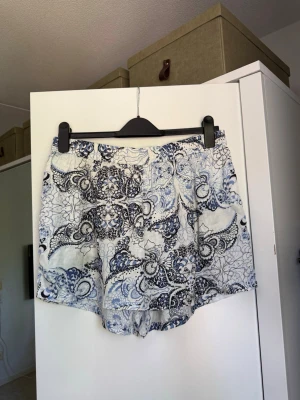 Blåvita shorts med paisleymönster - Säljer ett par blåvita shorts med paisleymönster. De har en loose passform och är tillverkade i ett tunt, mjukt material som känns skönt mot huden. Perfekta för varma sommardagar och har elastisk midja för extra komfort. Från märket Odd Molly i storlek 3 som enligt deras storlekguide motsvarar en storlek L. Mycket fint skick, jag hittar inga synliga anmärkningar eller defekter. 