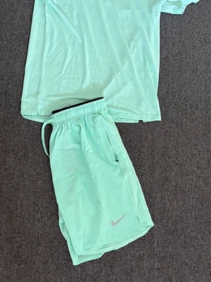 Mintgrönt träningsset från Nike - Snyggt mintgrönt träningsset från Nike med t-shirt och shorts. Båda plaggen har Nike-logga i silver och är tillverkade i lätt, snabbtorkande material som passar perfekt för löpning eller gym. T-shirten har rund hals och shortsen har dragsko i midjan. Storlek Xl men passar även L