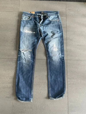 Dondup jeans - Säljer ett par blå jeans med raka ben och slitningar på både fram- och baksida. Jeansen har klassisk femficksdesign, ljusa tvättade partier och trasiga detaljer vid knät och låret. Perfekta för en avslappnad och trendig look.