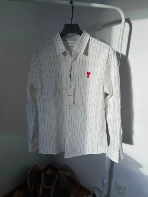 White Ami Shirt - Säljer en vit skjorta med tunna svarta ränder och ett broderat rött hjärta på bröstet. Skjortan har klassisk krage, långa ärmar och knäppning framtill. Perfekt för dig som gillar stilrena detaljer och enkel design.
