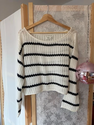 Vit stickad tröja med svarta ränder - Snygg vit stickad tröja med svarta horisontella ränder och bred halsringning. Tröjan har en avslappnad passform och långa ärmar. Perfekt för en trendig och avslappnad look.