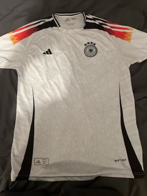 Tysklands matchtröja Adidas vit - Säljer en vit Tyskland matchtröja från Adidas med svarta detaljer och mönster i tyget. Tröjan har färgglada ränder i rött, orange och gult på axlarna, samt Tysklands landslagsemblem och Adidas-logga på bröstet. Tillverkad i lätt och ventilerande material.
