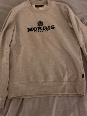 Beige sweatshirt från Morris  - Snygg beige sweatshirt från Morris  med svart broderad logga och emblem på bröstet. Tröjan har rund halsringning, långa ärmar och ribbade muddar vid ärmslut och nederkant. Perfekt för en avslappnad och stilren look.