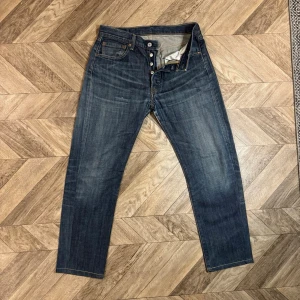 Mörkblå 501 Levi's - Säljer ett par klassiska mörkblå jeans från Levi's med raka ben och fem fickor. Jeansen har knappgylf och tydliga slitningar för en cool vintage-look. Perfekta för dig som gillar tidlös denimstil.