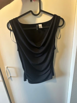 Svart draperad ärmlös topp - Snygg svart topp med draperad framsida och djupare ringning. Ärmlös modell i mjukt, stretchigt material som ger en smickrande passform. Perfekt att styla med jeans eller kjol för en trendig look.
