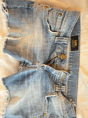 Ljusblå jeansshorts från Lee - Säljer ett par ljusblå jeansshorts från Lee med råa, fransiga kanter och klassisk femficksdesign. Shortsen har gylf med dragkedja och knapp, samt Lee-logga på baksidan, storlek xs
