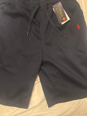 Marinblå shorts från Polo Ralph Lauren - Snygga marinblå shorts från Polo Ralph Lauren med elastisk midja och snörning. Klassisk röd logga broderad på sidan. Perfekta för sommaren och har en avslappnad passform. Tillverkade i mjukt material för extra komfort.
