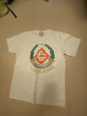 Vit t-shirt med Casablanca-tryck - Vit t-shirt med färgglatt Casablanca-tryck på bröstet. Trycket har ett emblem med gröna lagerblad, röd diamantform och texten 'Casablanca' i gult. Klassisk passform och rund halsringning. Perfekt för dig som gillar streetwear och snygga prints.