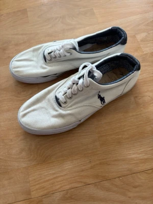 Vita sneakers från Polo Ralph Lauren - Snygga vita sneakers i canvas från Polo Ralph Lauren med klassisk låg profil och mörkblå logga på sidan. Skorna har vita snören och en vit sula, samt mörkblå detaljer på insidan. Perfekta för en clean och stilren look.