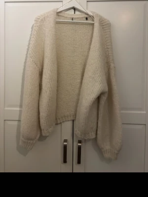 Oversized stickad kofta i offwhite - Mysig oversized stickad kofta i offwhite med grovstickad struktur. Koftan har långa ärmar med ribbade muddar och öppen front utan knäppning. Perfekt att slänga på sig för en avslappnad look.