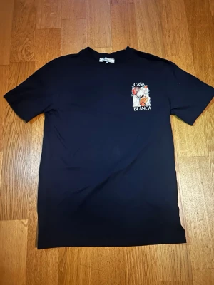 Marinblå Casablanca märke. Storlek M men passar även S - Svart t-shirt med färgglatt tryck och texten 'Casa Blanca' både fram och bak. Trycket har motiv av pelare, kortlek och palett. Klassisk passform och rund hals. Perfekt för dig som gillar unika prints.