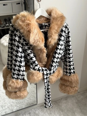 Houndstooth pälsjacka med bälte - Säljer en svartvit houndstooth-mönstrad pälsjacka med stora beige fuskpälskanter på krage, ärmslut och midja. Jackan har ett matchande bälte i midjan och en oversized look. Perfekt för dig som vill sticka ut i vinter!