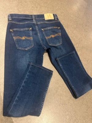 Mörkblå jeans från Nudie Jeans - Snygga mörkblå jeans från Nudie Jeans med klassisk femficksmodell och gul kontrastsöm. Bakfickorna har den ikoniska vågiga sömmen. Jeansen har rak/slim passform och är tillverkade i ett mjukt denimtyg som sitter skönt hela dagen. Är i storlek 29/32 men uppsydda på Jeansbolsget så är strl 29/30. Nudie jeans Grim Tim indigo myth. Nypris ungefär 1600:- 