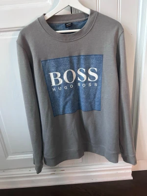 Grå sweatshirt från Hugo Boss - Snygg grå sweatshirt från Hugo Boss med stort blått tryck och vit logga på bröstet. Tröjan har rund halsringning och långa ärmar. Perfekt för en avslappnad och trendig look.