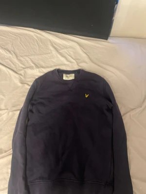 Mörkblå sweatshirt från Lyle & Scott - En stilren mörkblå sweatshirt från Lyle & Scott i ekologisk bomull. Tröjan har rund halsringning, långa ärmar och den klassiska gula loggan broderad på bröstet. Perfekt för en avslappnad och snygg look.