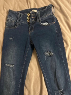 Mörkblå ripped jeans med låg midja  - Snygga mörkblå jeans med  låg midja och fyra dekorativa knappar framtill. Jeansen har slitna detaljer och hål på benen för en trendig look. Klassisk femficksmodell i stretchigt denimtyg som sitter tajt och bekvämt. Passar dig om du är kortare en 158 och inte längre en 162 då de är fult uppsydda därav priset 
