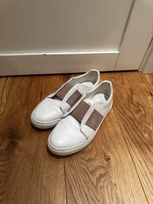 Vita sneakers från Philip Hog - Stilrena vita sneakers från Philip Hog med breda beige resårband över vristen. Skorna är låga och har en minimalistisk design i slätt läder med rund tå. Perfekta för en clean och modern look.