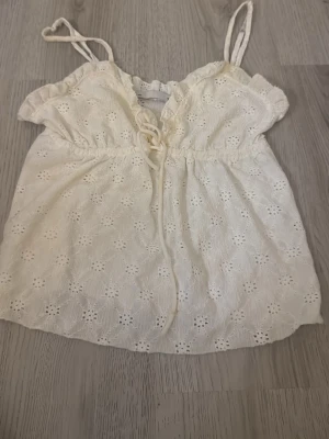 Vit broderad linnetopp med snörning - Söt vit linnetopp med tunna axelband, broderade blommor och snörning framtill. Toppen har en lätt volangkant upptill och är perfekt för varma dagar. Luftig och feminin design med fina detaljer.