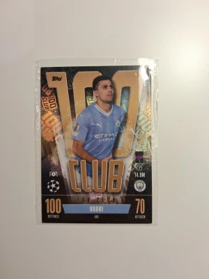 Rodri 100 Club Match Attax kort - Match Attax samlarkort med Rodri från Manchester City. Kortet är en 100 Club special med guldiga och svarta detaljer, Rodri i ljusblå tröja och klubbens logga. Perfekt för dig som gillar fotboll och samlarkort.