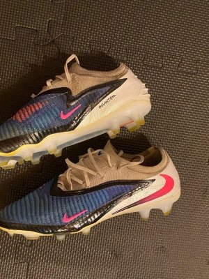 Nike Phantom elite blå och rosa fotbollsskor - Säljer ett par Nike Phantom fotbollsskor i beige, blå och svart med rosa detaljer. Skorna har en låg profil, snörning och mönstrad ovandel för bra bollkontroll. Sulan är gjord för spel på gräs och har gula dobbar. Perfekta för dig som vill sticka ut på planen.