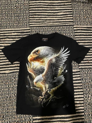 Svart t-shirt med mörkerlysande örntryck - Cool svart t-shirt med ett stort och detaljerat mörkerlysande tryck av en örn på bröstet och ryggen. Motivet har både en flygande örn och ett närbildsporträtt, vilket ger en riktigt kraftfull look. T-shirten har korta ärmar och rund hals, perfekt för dig som gillar djurmotiv.