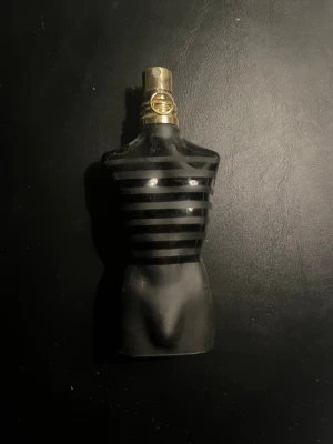 Svart parfymflaska i torsoform - Unik parfymflaska formad som en manlig torso i svart glas med horisontella ränder och guldfärgat sprayhuvud. Flaskan har en stilren och modern design som sticker ut på hyllan och passar perfekt för dig som gillar coola detaljer. Flaskan är lite mer än halvfull.