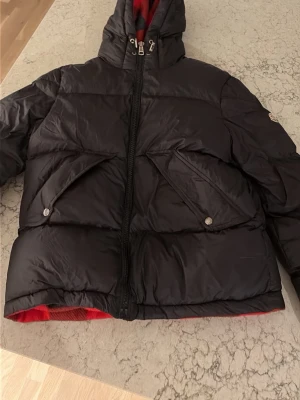 Svart pufferjacka från Moncler - Säljer en svart pufferjacka från Moncler med stor huva och röda detaljer på insidan. Jackan har två stora fickor framtill med lock och tryckknappar samt dragkedja hela vägen upp. Perfekt för kalla vinterdagar och riktigt snygg streetstil.
