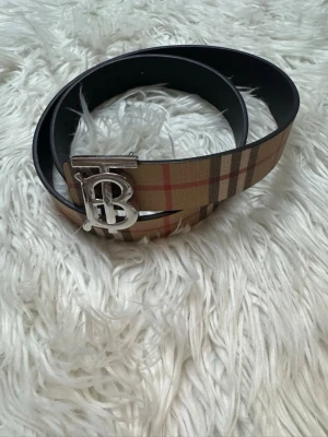 Rutig Burberry bälte med silverdetalj - Snyggt bälte från Burberry med klassiskt rutig design i beige, brunt, svart och rött. Bältet har en stor silverfärgad spänne med Burberrys ikoniska TB-logga. Perfekt för att lyfta din outfit med en lyxig touch.