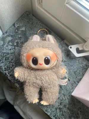 Söt beige labubu nyckelring PRIS KAN DISKUTERAS! - Huvudet sitter lite på sneddan! Nyckelring med en gullig labubu i beige fluffig plyschdräkt med kaninöron. Stora svarta ögon och orangea kinder som ger ett charmigt uttryck. Perfekt att hänga på väskan eller nycklarna för en personlig touch. Ursnyggt till hösten!