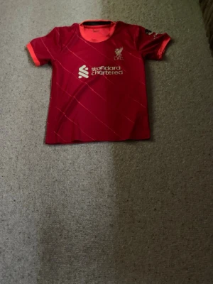 Liverpool matchtröja Firmino #9 - Röd Liverpool FC matchtröja med vita detaljer och tryck av sponsor på bröstet. På ryggen står det Firmino och nummer 9. Tröjan har korta ärmar med orange muddar och rund hals. Perfekt för dig som älskar fotboll och vill stötta ditt lag. Passar personer som är på 140 cm +\- 10 cm. Ca 10 åring