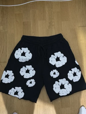 Svarta shorts med vita blommor - Säljer ett par svarta shorts med stora vita blommönster över hela plagget. Shortsen har resårmidja med snörning och är tillverkade i ett mjukt material som passar perfekt till sommaren. De har en loose passform och är riktigt sköna att bära.