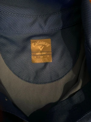Blå pikétröja från Callaway - Snygg blå pikétröja från Callaway med klassisk krage och knappar framtill. Tillverkad i ett lätt och funktionellt material som andas, perfekt för golf eller andra aktiviteter. Callaway-logga broderad på bröstet.