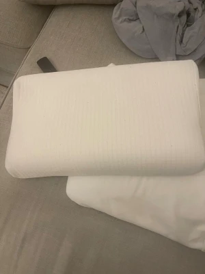 Vitt ergonomiskt memory foam-kudde - Säljer en vit ergonomisk kudde i memory foam med lätt böjd form för bättre nackstöd. Kudden har ett avtagbart tygöverdrag med diskret randigt mönster. Perfekt för dig som vill ha extra stöd när du sover eller vilar.