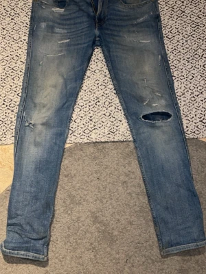 Ljusblåa jeans med hål från Replay - Säljer ett par ljusblå replay jeans med skinny fit och flera slitningar, inklusive ett större hål på ena benet. Jeansen har klassisk femficksmodell och är tillverkade i denim. Perfekta för en avslappnad och trendig look.