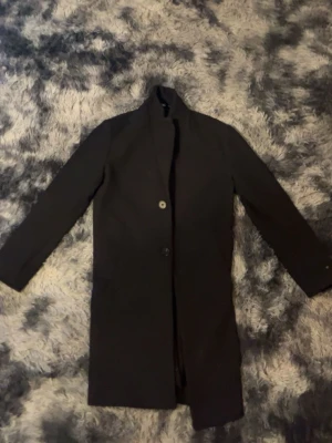 Svart klassisk rock med ståkrage - Snygg svart rock med ståkrage och två knappar framtill. Lång modell med raka ärmar och diskreta fickor på sidorna. Perfekt för dig som vill ha en stilren och tidlös look. Materialet ser ut att vara ullblandning.