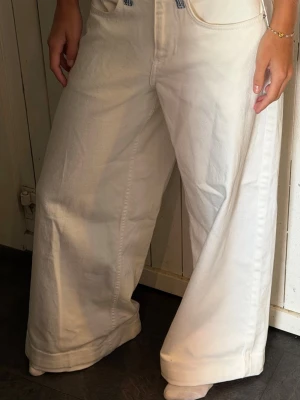 Vida vita jeans med blåa detaljer - Säljer ett par vita jeans med bred passform och raka ben. Jeansen har blå- och vitrandiga detaljer i midjan och bakfickor med lock och knapp. Perfekta för en avslappnad och trendig stil. Uppsydda ner till!