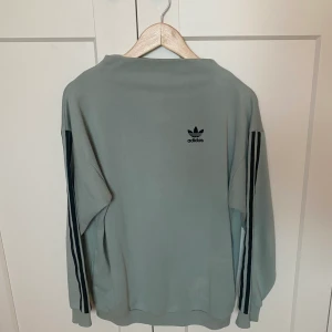 Adidas sweatshirt - En tjocktröja från Adidas i en pastellig grönblå färg med svarta detaljer. Tröjans halsöppning är något högre och bildar en lös polokrage.  Storlek 34 men modellen är oversized så beroende på hur löst sittande man vill ha den kan den passa flera storlekar.