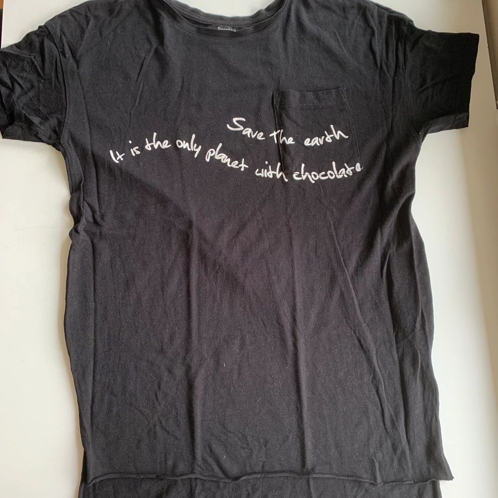 Svart t-shirt med en liten fick på vänster sida. På det vita trycket står det ”Save the earth It is the only planet with chocolate”. Längre tyg bak och en liten slits i sidan. Fint skick och super skönt material. Stl XS från Bershka. Tröjan koster 30 kr, frakt ingår ej. Swipe för fler bilder.. T-shirts.