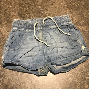 Blå tunna och lätta jeansshorts 34 - Blåa jeansshorts med rep i resorband runt midjan. Två vita knappar på vardera sida av benslut. Helt nya lite skrynkliga.